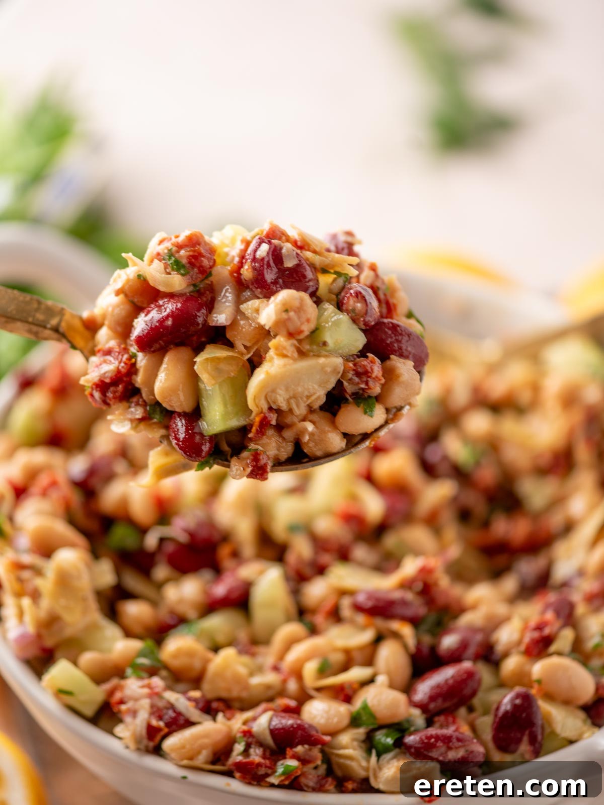 A spoonful of mixed bean salad with kidney beans, chickpeas, white beans, chopped cucumber, parsley, and sun-dried tomatoes, held above a bowl filled with the same salad.