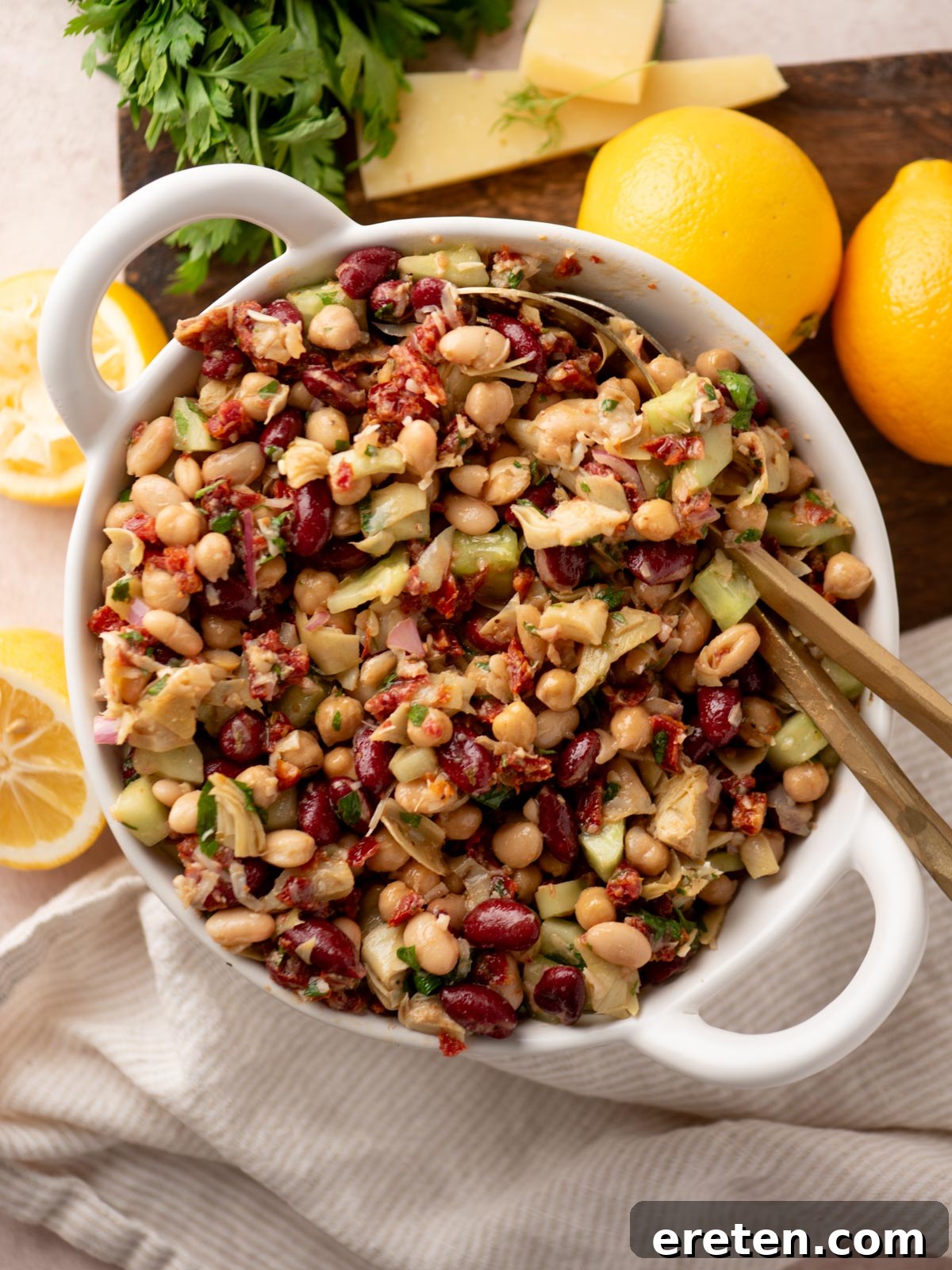 A white bowl filled with a colorful bean salad featuring kidney beans, chickpeas, white beans, chopped vegetables, and herbs. Lemons, cheese slices, and fresh parsley are on a wooden board in the background.