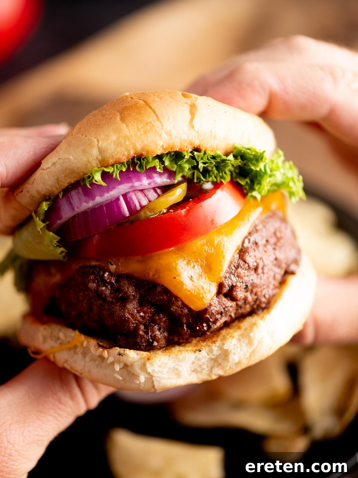 Juicy Grilled Burger Recipe: Charred, Flavor-Packed Patties 2 A close-up of hands holding a hamburger with a beef patty, melted cheddar cheese, tomato, red onion, pickles, and lettuce in a sesame seed bun.