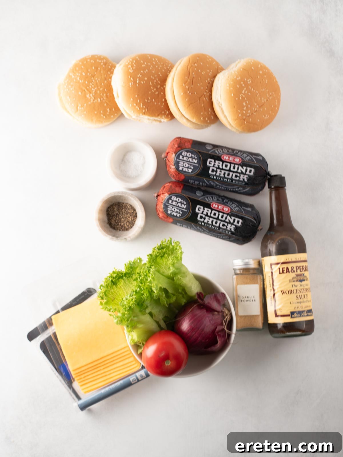 Juicy Grilled Burger Recipe: Charred, Flavor-Packed Patties 3 Top-down view of burger ingredients: four sesame seed buns, two rolls of ground chuck, salt, pepper, Worcestershire sauce, garlic powder, cheese slices, lettuce, a tomato, and a red onion on a white surface.