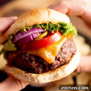 Juicy Grilled Burger Recipe: Charred, Flavor-Packed Patties 6 A close-up of hands holding a cheeseburger with a beef patty, melted cheddar cheese, tomato slices, red onion, pickles, and lettuce in a bun.