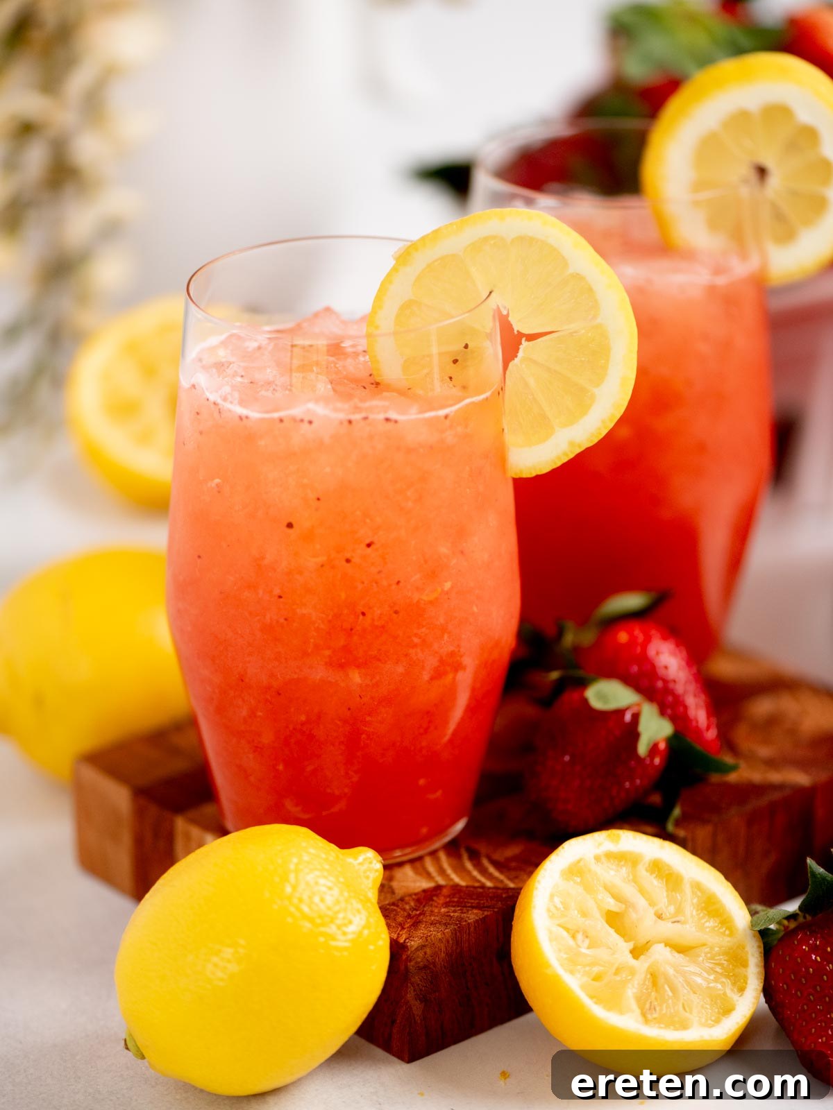 Two glasses of strawberry lemonade with lemon slices on the rims, surrounded by fresh strawberries and lemons, including a halved lemon, on a wooden board. The bright drinks look refreshing and vibrant.
