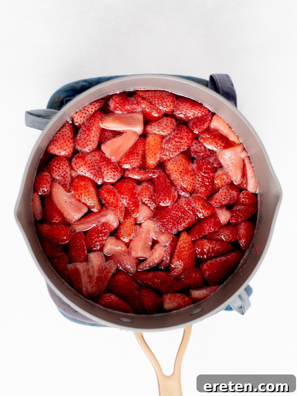 A pot filled with chopped strawberries sits on a blue oven mitt, viewed from above. The strawberries are bright red and appear to be cooking.