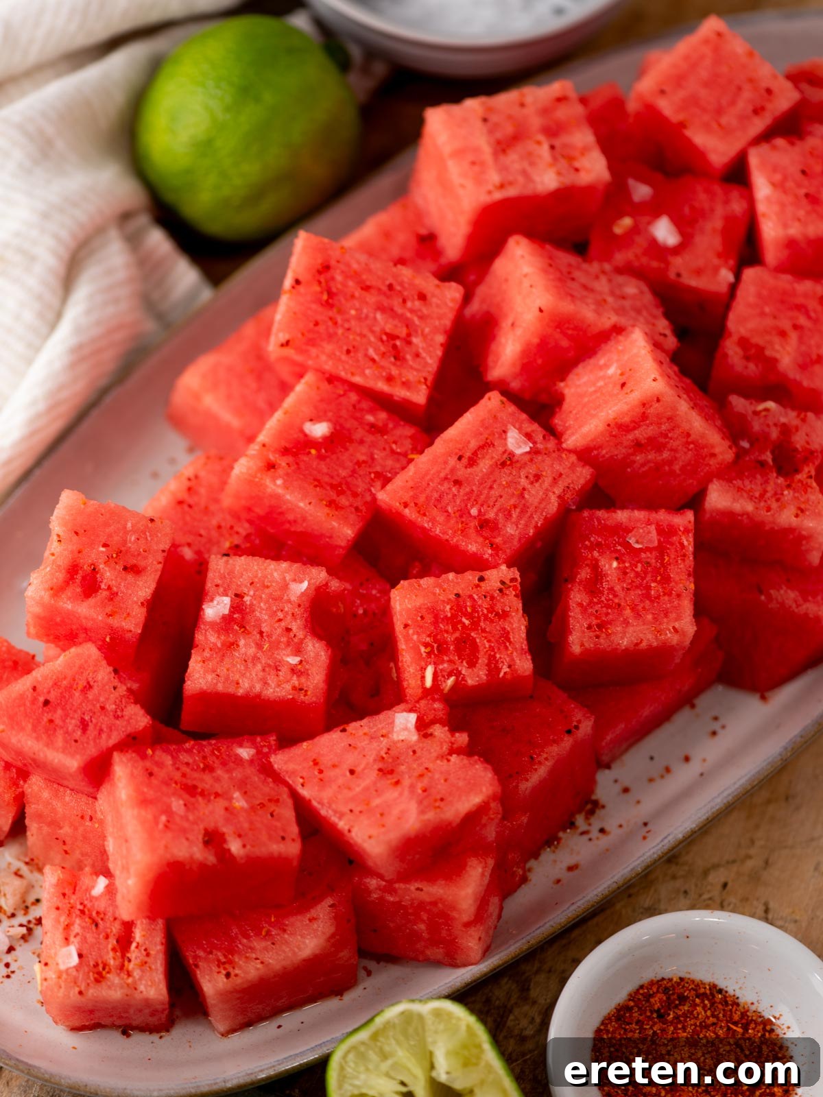 Watermelon with Tajín: Spicy Lime and Chili Seasoning 2 A platter of cubed watermelon sprinkled with chili powder and salt, surrounded by lime and chili seasoning bowls on a wooden surface.