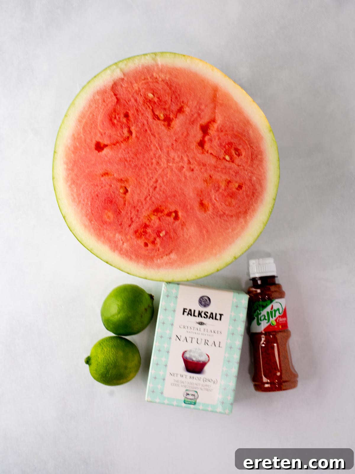 Watermelon with Tajín: Spicy Lime and Chili Seasoning 3 A halved watermelon, two limes, a box of Falksalt natural salt, and a bottle of Tajin seasoning are arranged on a white surface.