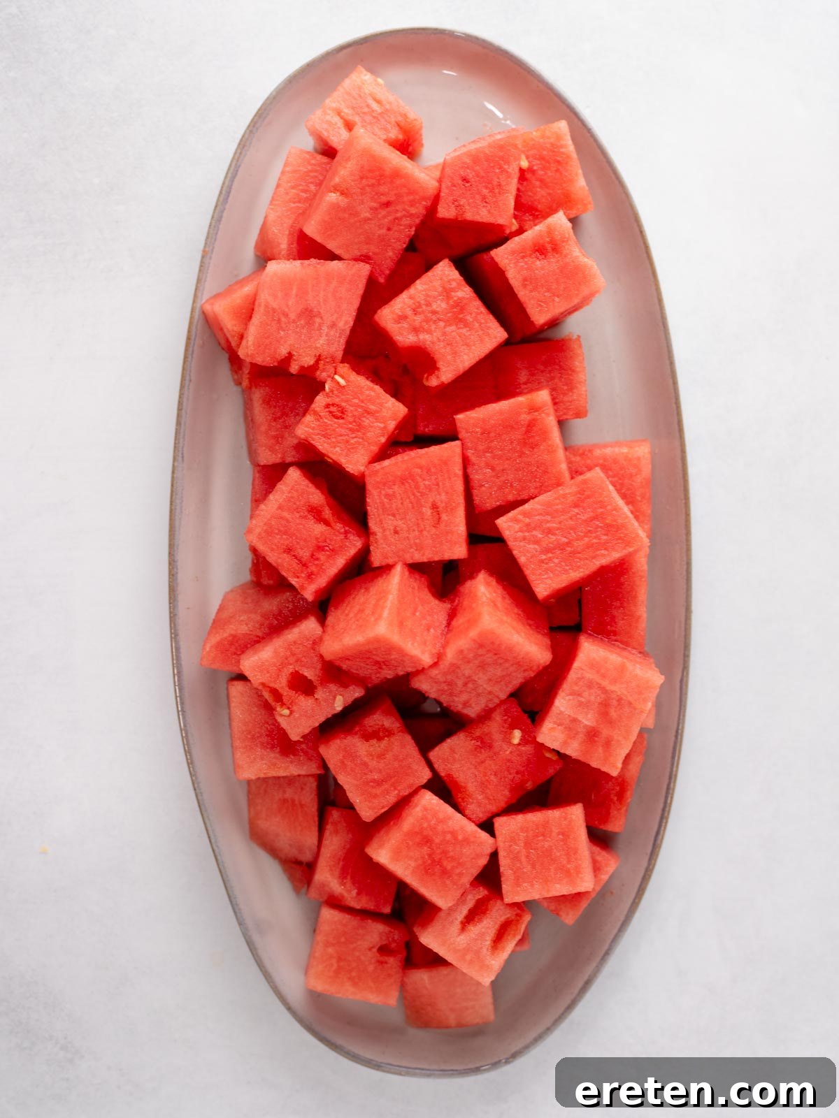 Watermelon with Tajín: Spicy Lime and Chili Seasoning 4 A clear oval plate filled with evenly cut cubes of fresh, red watermelon, placed on a light-colored surface.