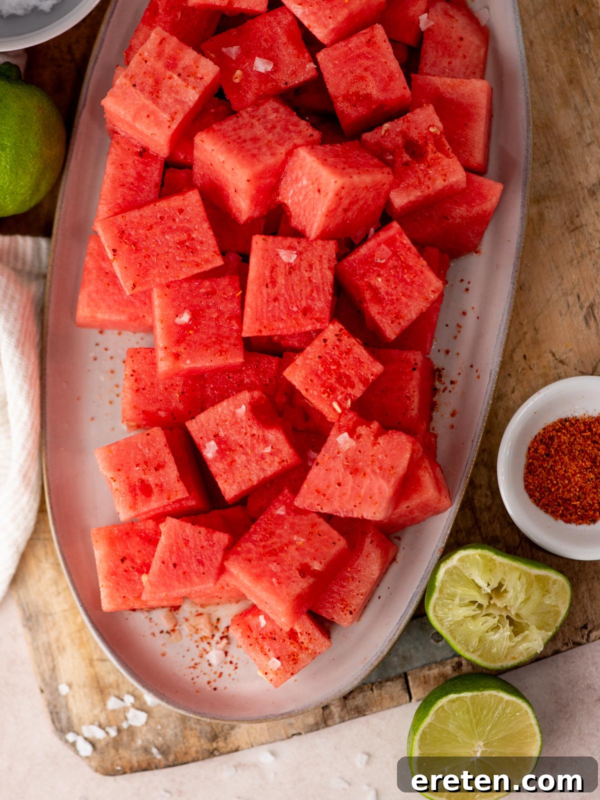 Watermelon with Tajín: Spicy Lime and Chili Seasoning 5 Cubed watermelon sprinkled with chili powder and coarse salt on a platter, surrounded by halved limes, a bowl of chili powder, and a wooden surface.